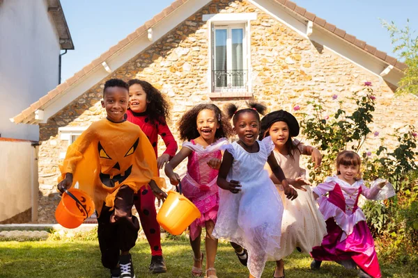 一群身穿万圣节服装的孩子们穿着紧身衣在草坪上跑着 他们一起在屋前用糖果桶尖叫着 — 图库照片