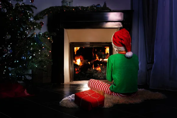 Boy from back with Christmas present in New Year costume sit near fireplace and looking on fire