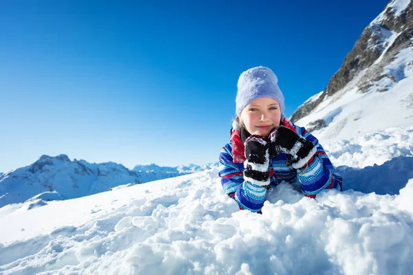 快乐微笑的小女孩躺在雪地里 双手抱头 头戴冬帽 背对着山 — 图库照片