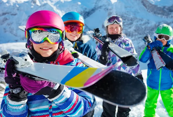 幸せな笑顔若いです女の子でピンクヘルメットと色ゴーグルホールドスキーで彼女の手で友人のグループの子供たちにバックグラウンドで山 — ストック写真