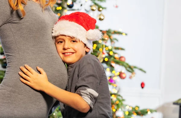 圣诞树前 妈妈和小男孩戴着圣诞老人帽子抱着妈妈的肚子的画像 — 图库照片