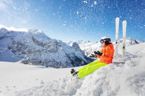 幸せな女の子でオレンジ明るいスポーツ衣装とともにスキースローアップ雪で空気中に座る上の高い山脈のピークと青空 — ストック写真