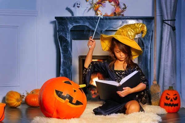 Halloween portrait of wizard girl sit with magic stick witch hat and spell book by fireplace