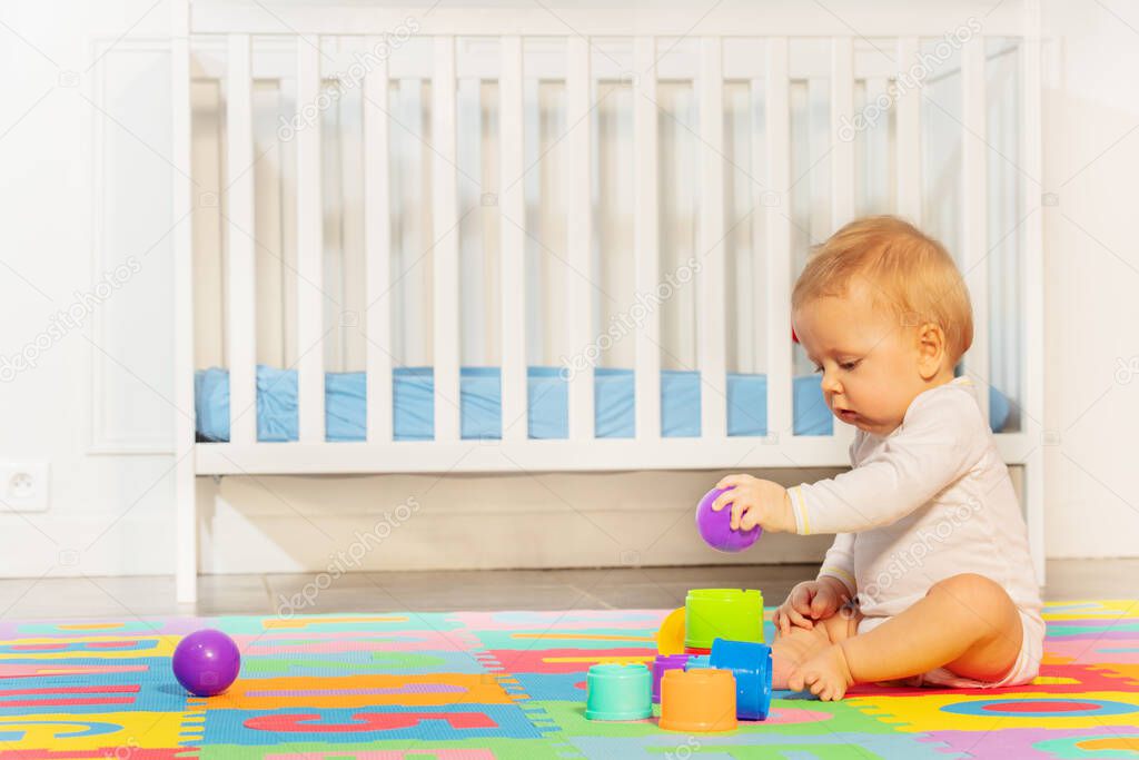 Little toddler baby boy sit in the room on children carpet near crib build pyramid of blocks