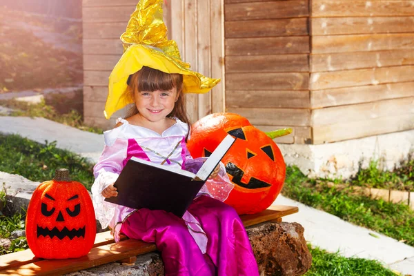 小さなハロウィン魔女女の子とともにマジック本座っています外でザベンチ保持カボチャ — ストック写真