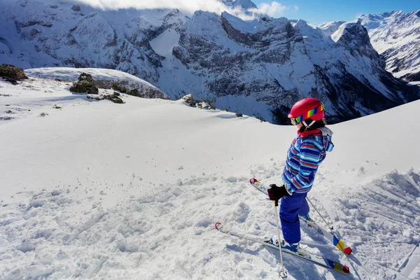 スキー場の女の子の上からの肖像画は 鮮やかなコートでピンクのヘルメットの後ろから山の景色の上に雪の中に立つ — ストック写真