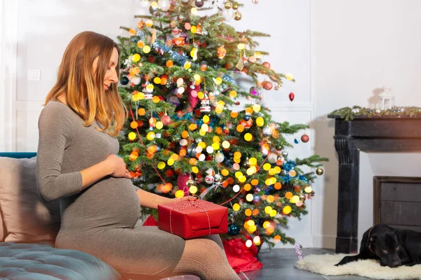 怀孕的女人摸着肚子 坐在壁炉边的圣诞树旁 拿着礼物坐在沙发上 — 图库照片