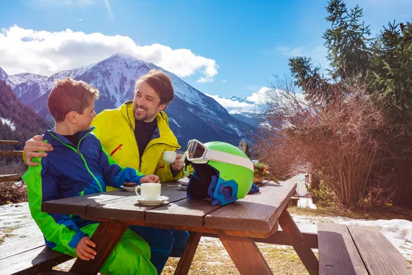 父亲抱着一个穿着滑雪服的小男孩 与他交谈 滑雪后享受山景下的午休 — 图库照片