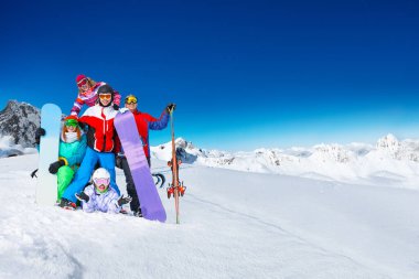 Yetişkin snowboardculardan oluşan bir grup sırt üstü güneşli havada, dağların tepesinde birlikte poz veriyorlar.