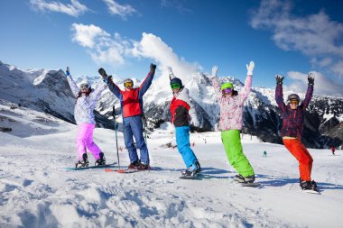 Kayak ve snowboarddaki genç yetişkinler el ele tutuşup dağların zirvesine çıkıyorlar.