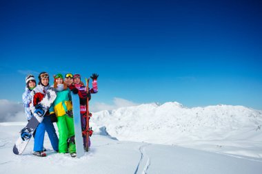 Yetişkin snowboardculardan oluşan bir grup sırt üstü güneşli havada, karla kaplı zirvelerin üzerinde birlikte dağın tepesinde dururlar.