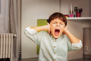 Little autistic boy with strong negative face expression closing ears and loudly screams clipart