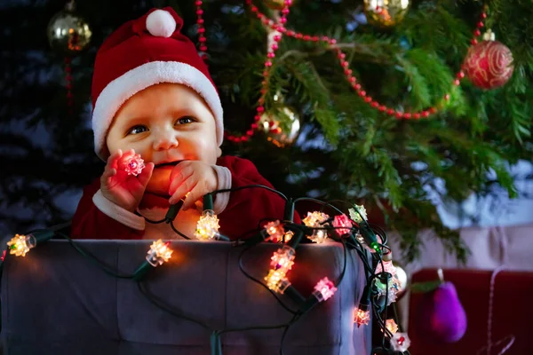 小可爱的小男孩 戴着圣诞老人的帽子 手里拿着灯火通明的灯 坐在圣诞树下的盒子里 看上去很可爱 — 图库照片