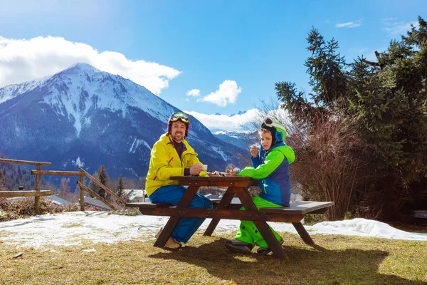 穿着滑雪服的快乐小男孩在高山全景上和父亲一起坐着吃午饭 — 图库照片
