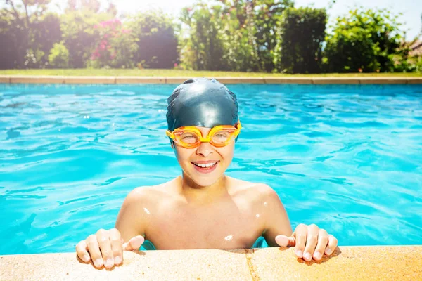 Cute little boy in swimming cap and googles lay on the border of outdoor pool look at camera smiling