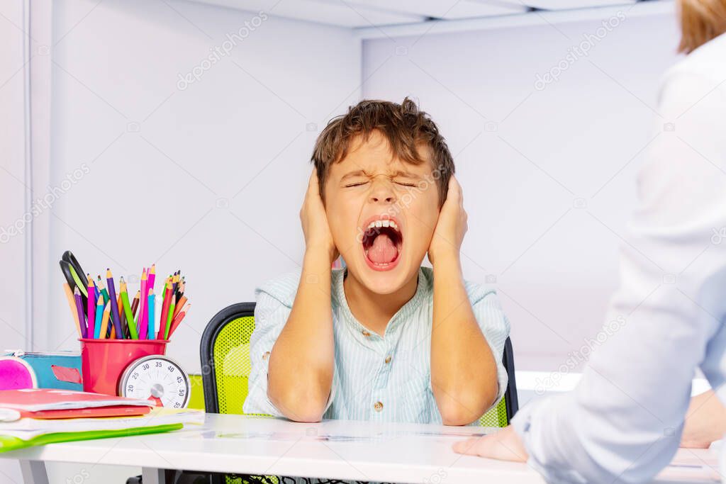 Boy with autism disorder scream and close ears in pain during ABA development therapy sitting by the table in class, sensory problem