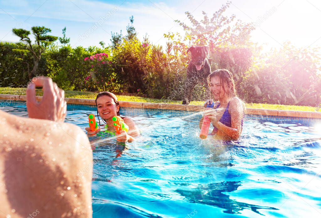 Portrait with two laughing girls in swimming pool shooting water-gun squirt pistol in boys on sunny day