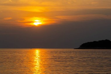 Sakin bir altın gün batımı, ufukta bir adanın karanlık silueti ve sakin denizi yansıtıyor. Seyahat, doğa ve rahatlama temaları için mükemmel..