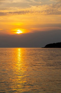 Sakin bir altın gün batımı, ufukta bir adanın karanlık silueti ve sakin denizi yansıtıyor. Seyahat, doğa ve rahatlama temaları için mükemmel..