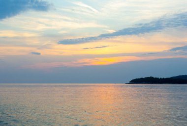 Sakin denizin üzerinde, sudaki yumuşak yansımaları ve ufukta karanlık bir ada silueti olan sakin, pastel renkli bir günbatımı. Seyahat, doğa ve rahatlama temaları için mükemmel..