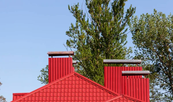 Red metal roof on the building. On the roof surface there are three chimneys closed with red metal tiles. Against the blue sky