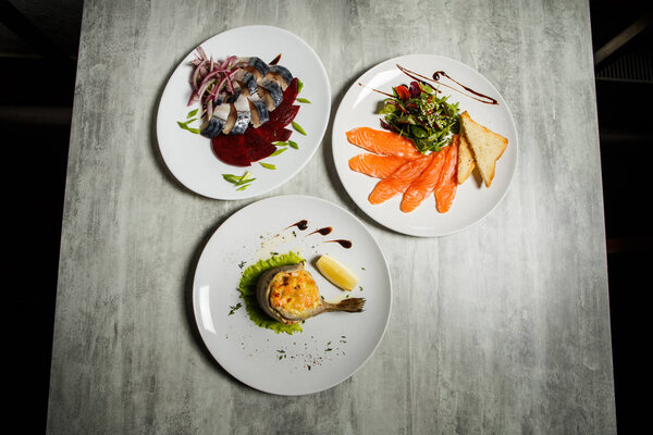 три разнообразных ресторанных блюда из скумбрии, лосося, форели и овощного гарнира с травами стоят на сером столе
