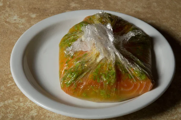 wrapped in food film piece of peeled raw salmon fillet marinated in lemon juice with chopped dill roots lie on white plate