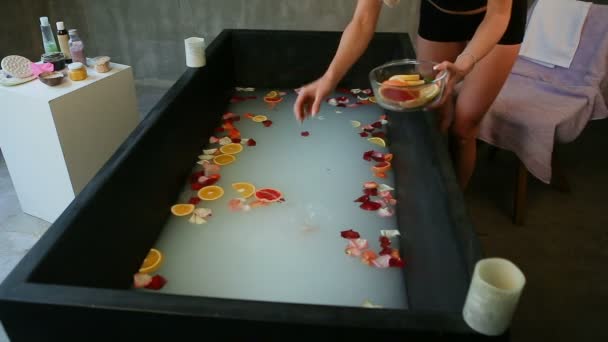 Тощая девушка бросает цитрусовые ломтики и лепестки роз в черную ванну — стоковое видео