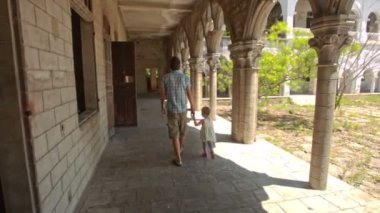 Baba küçük sarışın kız, beyaz gökyüzüne karşı sütunları olan eski gotik bina galerisinin gölgesinde çömelmiş.