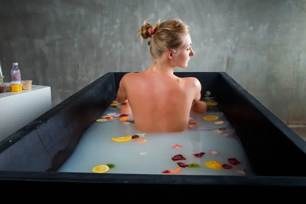 若い美しい金髪の女性が深い黒の浴槽でミルク カラフルなバラの花びら 柑橘類のスライスとお風呂に入る — ストック写真