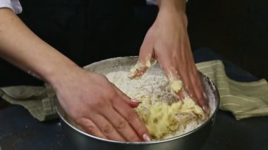 Ağır çekimde yakın plan kadın elleri beyaz unla yumuşak mayayı koyu metal kasede karıştırıyor. Mutfak masasında duruyor.