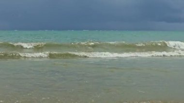 Mavi denizin güçlü beyaz dalgaları yaz mevsiminde kumlu gökyüzüne karşı yavaşça kumsala doğru koşuyor.