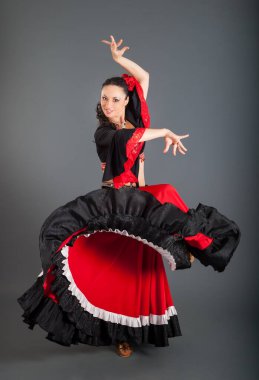 Kırmızı ve siyah çingene tarzı takım elbiseli genç esmer kadın ve saçlarında kırmızı güller stüdyoda gri arka planda dans ediyor.