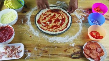 İnsan elinin üst görüntüsü kaşıkla pizza hamuruna domates sosu koyuyor.
