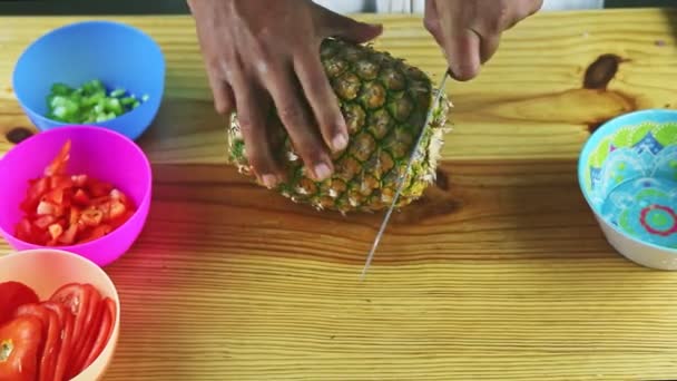 Shora pohled na ruce kuchaře nožem čistí ananas od slupek na dřevěném stole — Stock video