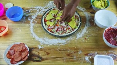Erkek ellerinin üst görüntüsü çiğ pizzaya farklı malzemelerle ananas dilimleri koyuyor.