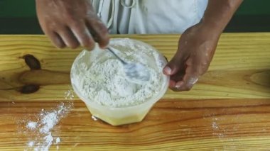 İnsan ellerinin üst görüntüsü kaşıkla karıştırılmış beyaz un ve yumuşak hamur plastik kasede.