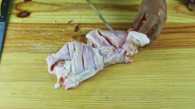 Üst görüntü insan eli, bıçakla, çiğ tavuğun yarısını ahşap masada kesiyor.