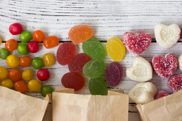 Sweets in paper bags. Multicolored candy, marmalade in the form of heart on a white wooden background