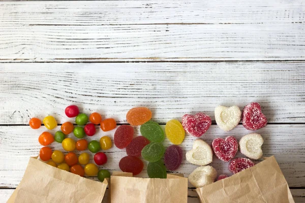 Sweets in paper bags. Multicolored candy, marmalade in the form of heart on a white wooden background