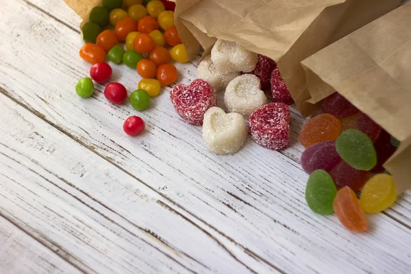 Sweets in paper bags. Multicolored candy, marmalade in the form of heart on a white wooden background