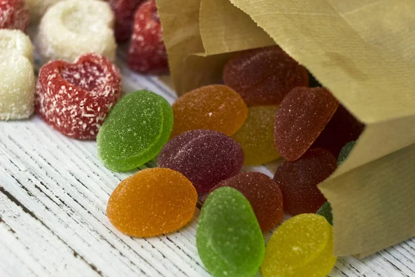 Round colored candy and marmalade in a paper bag on a wooden table, bright sweets, white background