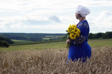 İşlemeli süslemeli mavi elbiseli genç bir kadın buğdaylı bir tarlanın ortasında bir buket ayçiçeği tutuyor. Ulusal giyinmiş Ukraynalı kadın sahada