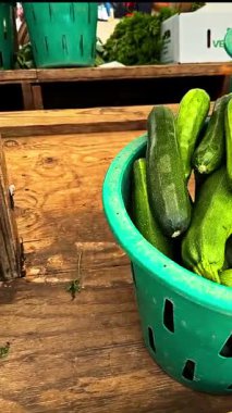 Shoppers enjoy browsing vibrant zucchinis in a green basket at a market. The atmosphere is lively, showcasing fresh local produce on a sunny day.