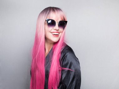 Uzun pembe saçlı beyaz bir kadın, güneş gözlüğü takıyor, yan bakıyor, gülümsüyor, gri arka planda soyutlanmış.