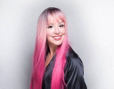 Uzun pembe saçlı beyaz bir kadın, ipek ev bornozu giyiyor, düz görünüyor, gülümsüyor, gri arka planda soyutlanmış.