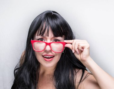 Beyaz tenli, siyah sağlıklı saçlı, beyaz tenli, kırmızı gözlüklü beyaz güzel kadının yakın plan portresi, gri arka planda gözlüklerine dokunuyor.