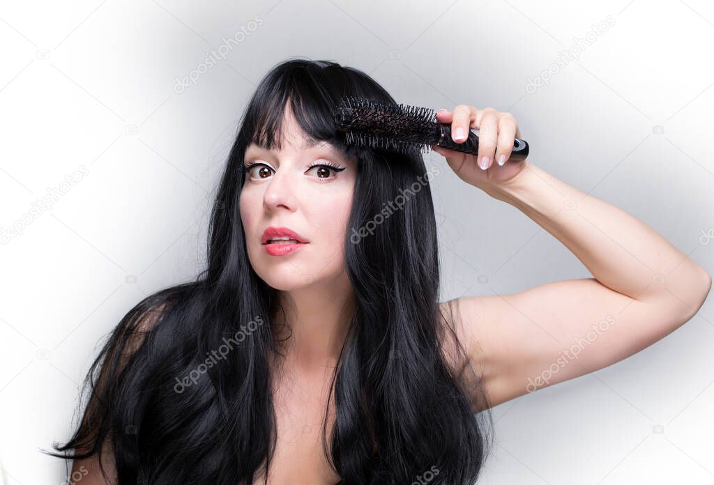 A brunette young woman is focused on brushing her hair with the brush, isolated on grey and white background