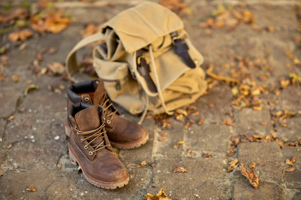 Brown leather boots and a canvas backpack stand on the floor with autumn dry leaves. Hikind main clothes