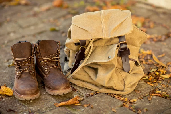 Brown leather boots and a canvas backpack stand on the floor with autumn dry leaves. Hikind main clothes